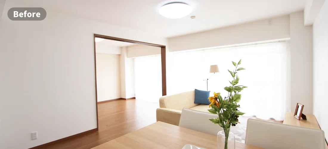 家具消しのステージングビフォー画像　生活感のあるリビングルーム。壁には絵が飾られており、ローチェスト、テレビ、本棚が壁沿いに、部屋中央にはローテーブルとソファがおかれ、窓にはベージュのカーテンがかかっている