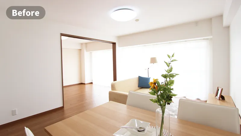 家具消しのステージングビフォー画像　生活感のあるリビングルーム。壁には絵が飾られており、ローチェスト、テレビ、本棚が壁沿いに、部屋中央にはローテーブルとソファがおかれ、窓にはベージュのカーテンがかかっている